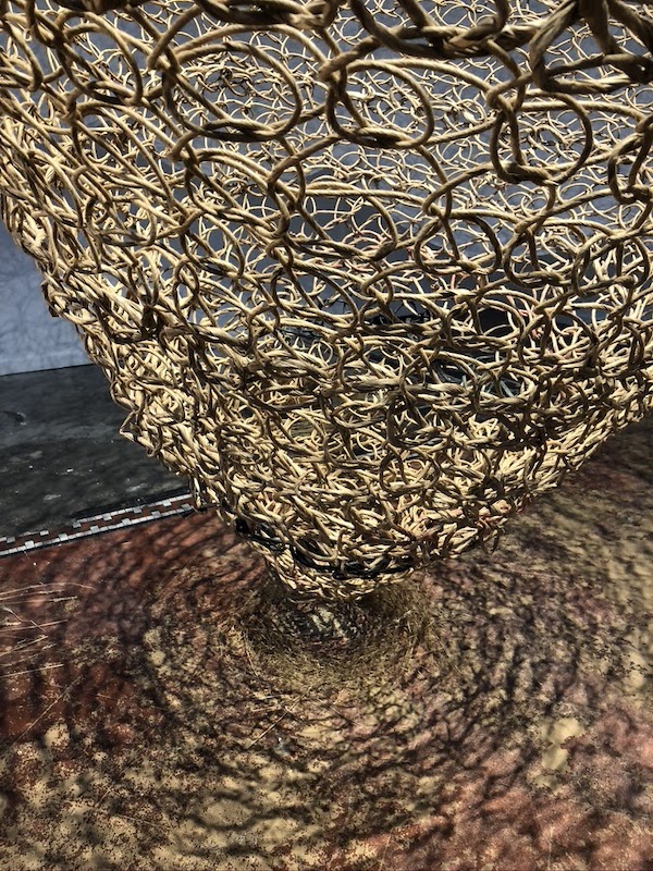 Rings of jute intertwine to form a massive trap-like net, textured by light and shadow that is reflected on the red, white, and grey tiled floor littered with dried grass.