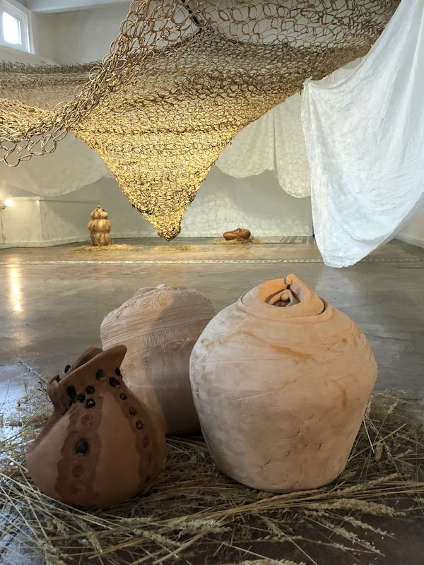 White linen draped from walls and ceiling, partially frames a mass of jute netting, which tapers down toward the red/brown tiled floor. On the floor sit several clay vessels nestled in straw, the shapes of which resemble female anatomy. Light shines in from a window and up from fixtures.