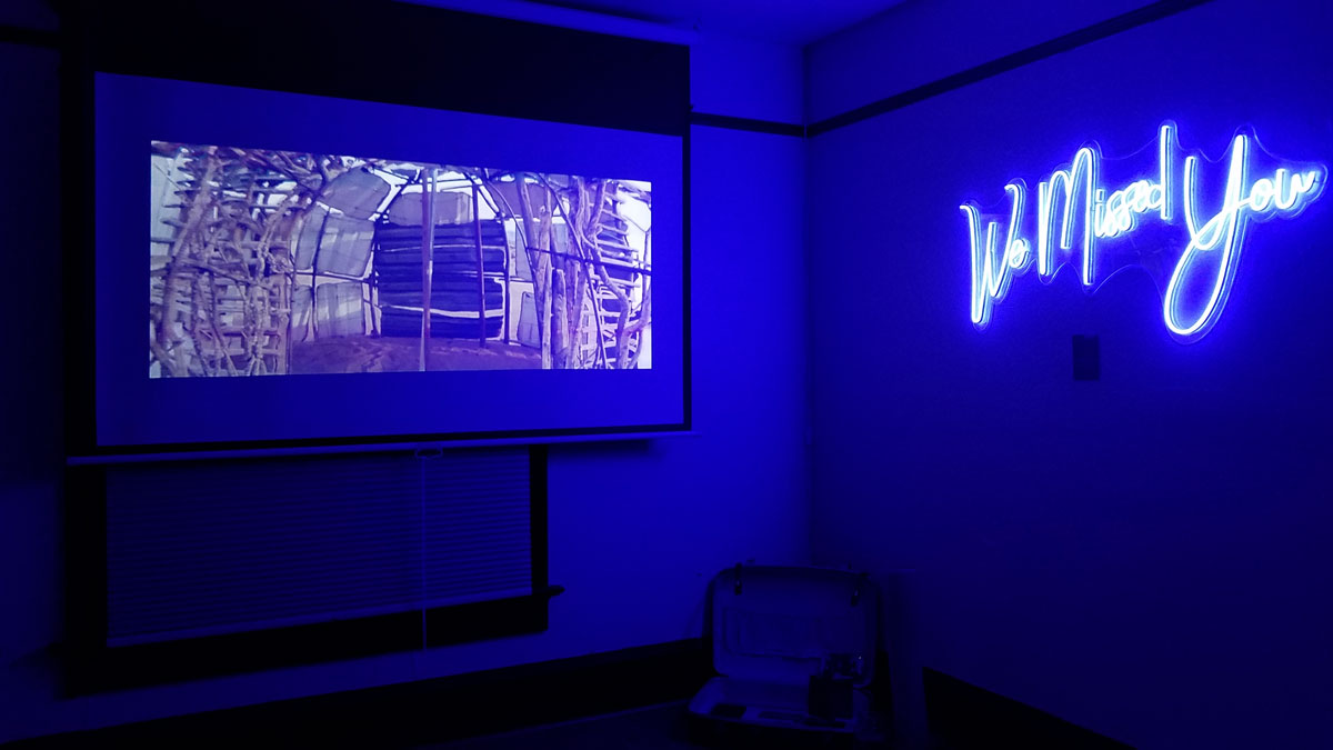 A room bathed in blue light has a video projection on one wall. On an adjoining wall, cursive neon letters spell ‘We Missed You’.