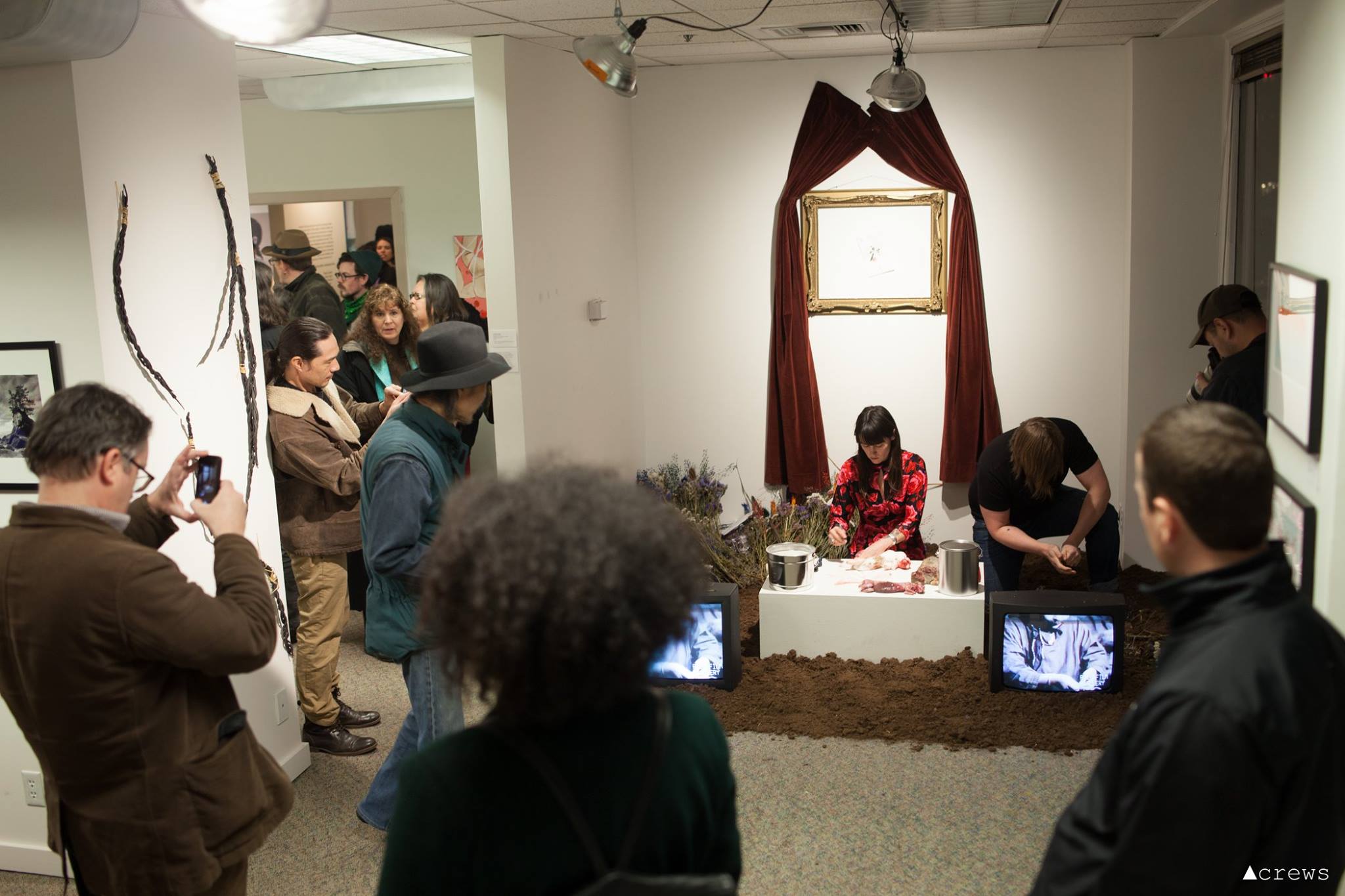 A crowd watches two performers in an art gallery. There is dirt on the floor and video monitors flanking a white pedestal with metal buckets and skinned rabbits. Behind, a gold frame on the wall is draped with red velvet curtains.