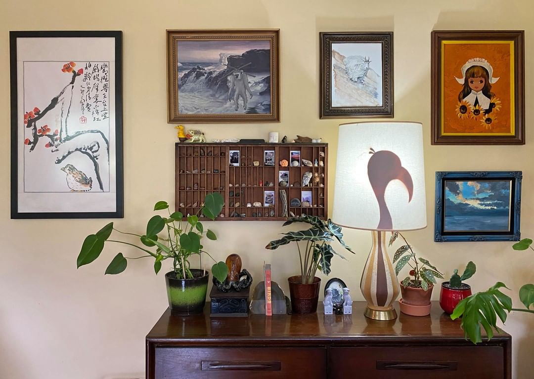 A variety of framed paintings and an old typesetter case with little objects hang on a wall above a chest of drawers with house plants, small sculptures and a bird-shaped lamp on top.