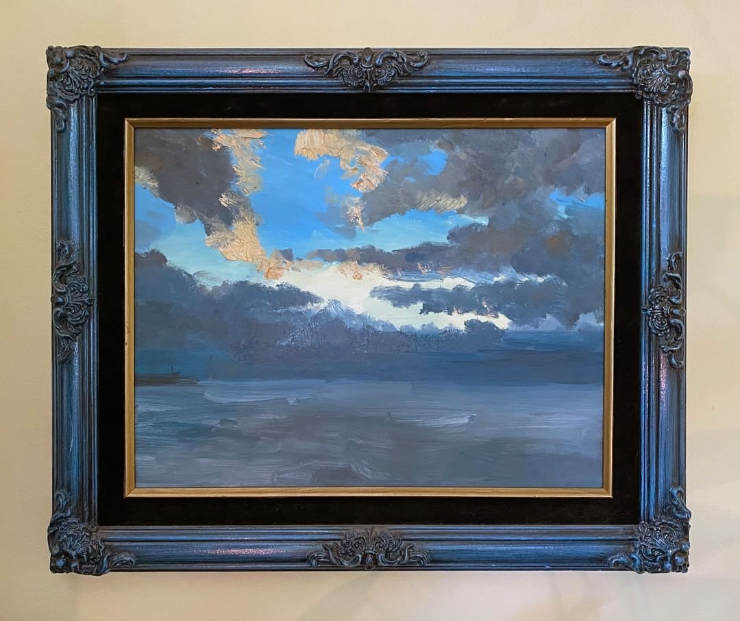 A seascape painting in an old-fashioned wood frame depicting a dramatic cloud-heavy sky with light breaking through.
