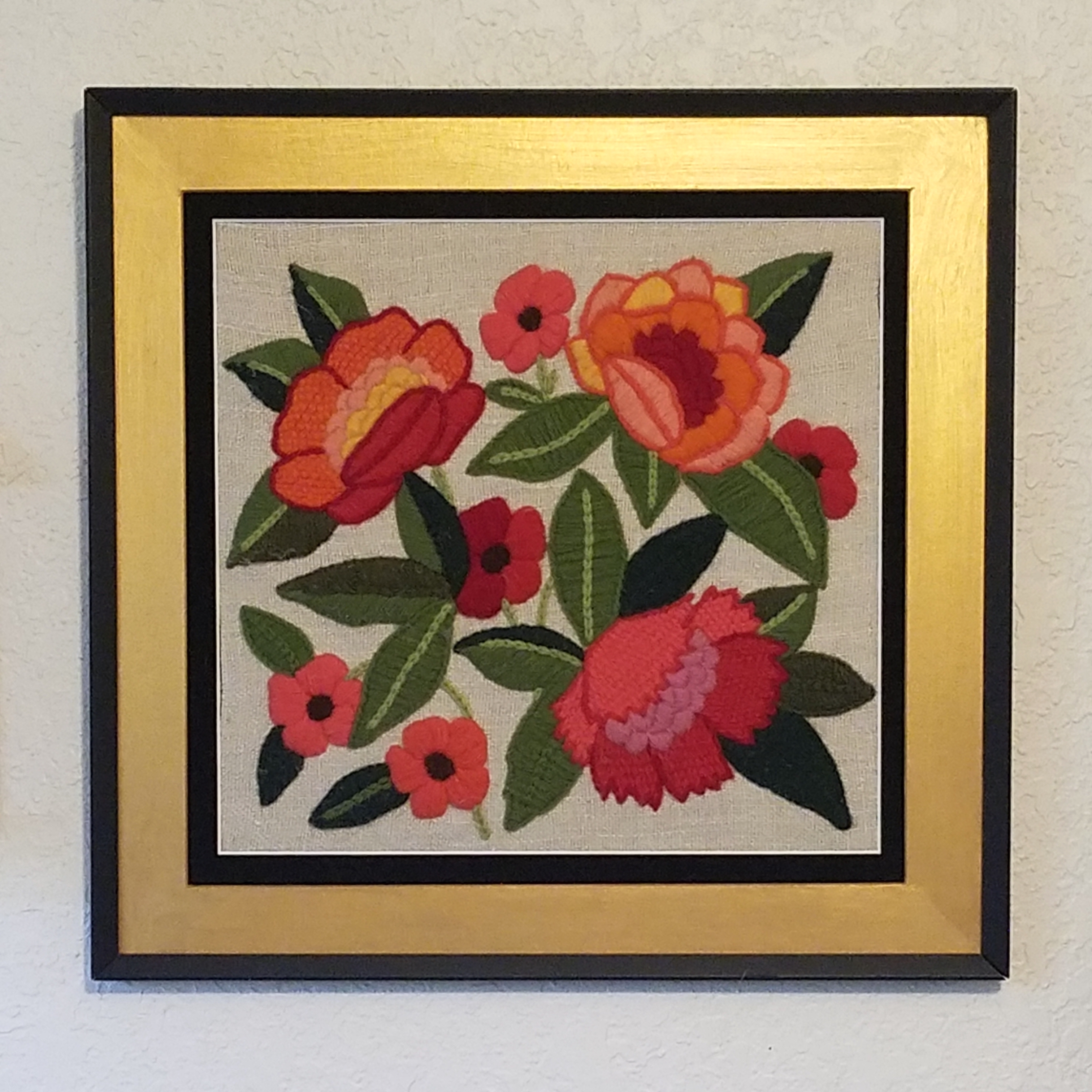 Three large and five small flowers in predominantly red and orange colors are embroidered on tan fabric, with a gold and black frame.