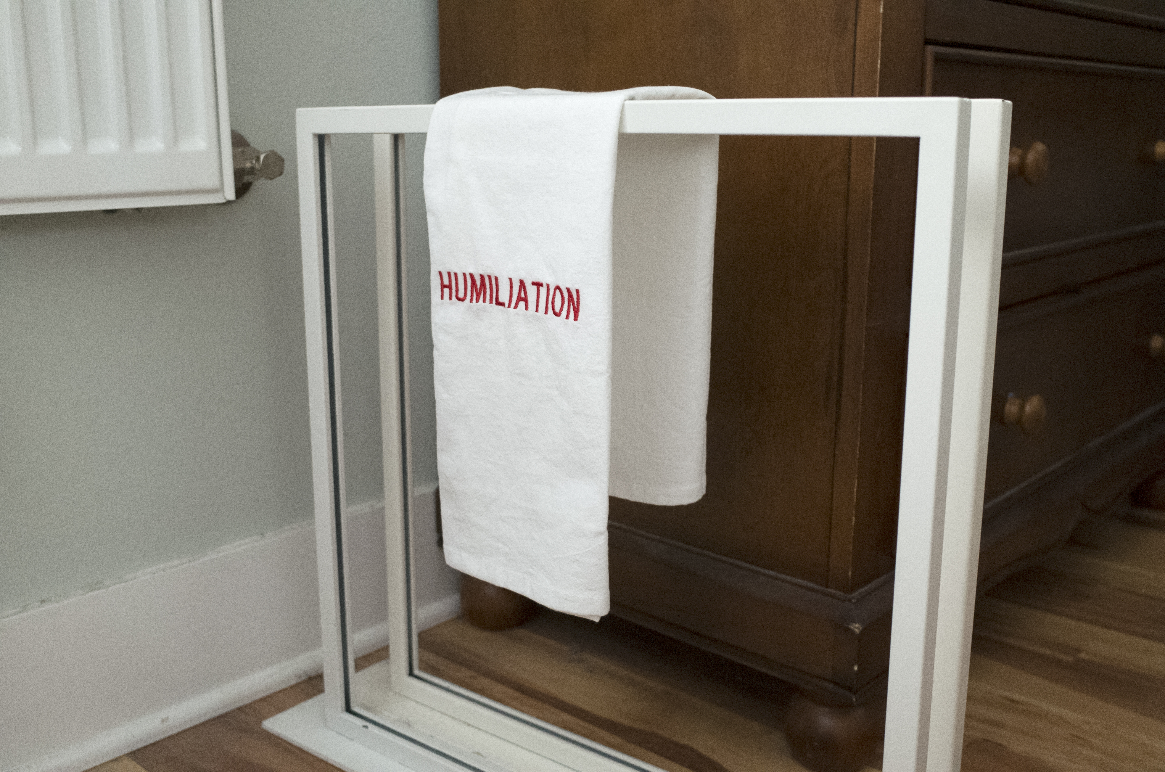 Two white metal frames stand upright and parallel to each other with a small gap in between on the floor of a domestic space. A folded white towel with the word ‘humiliation’ in red embroidery is draped over the top of the two frames.