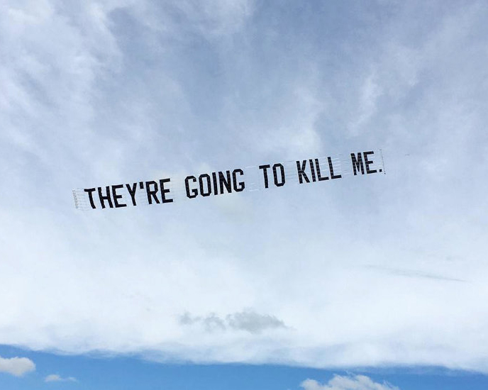 Large black capital letters being pulled across the sky by an airplane say “They’re going to kill me.” The sky is cloudy with a strip of blue at the bottom.