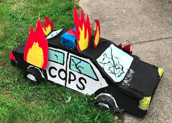 A black and white sedan made of painted cardboard has the word ‘COPS’ written across its side. Its windows are cracked and bright red and yellow cardboard flames shoot out of its roof and tail.