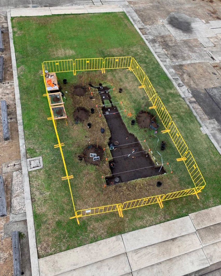 Overhead view of a grassy lawn with an excavated portion in the shape of a monumental statue on a pedestal. A bright yellow fence surrounds the area which also contains worktables, digging tools, signs and rectangular string boundary lines.