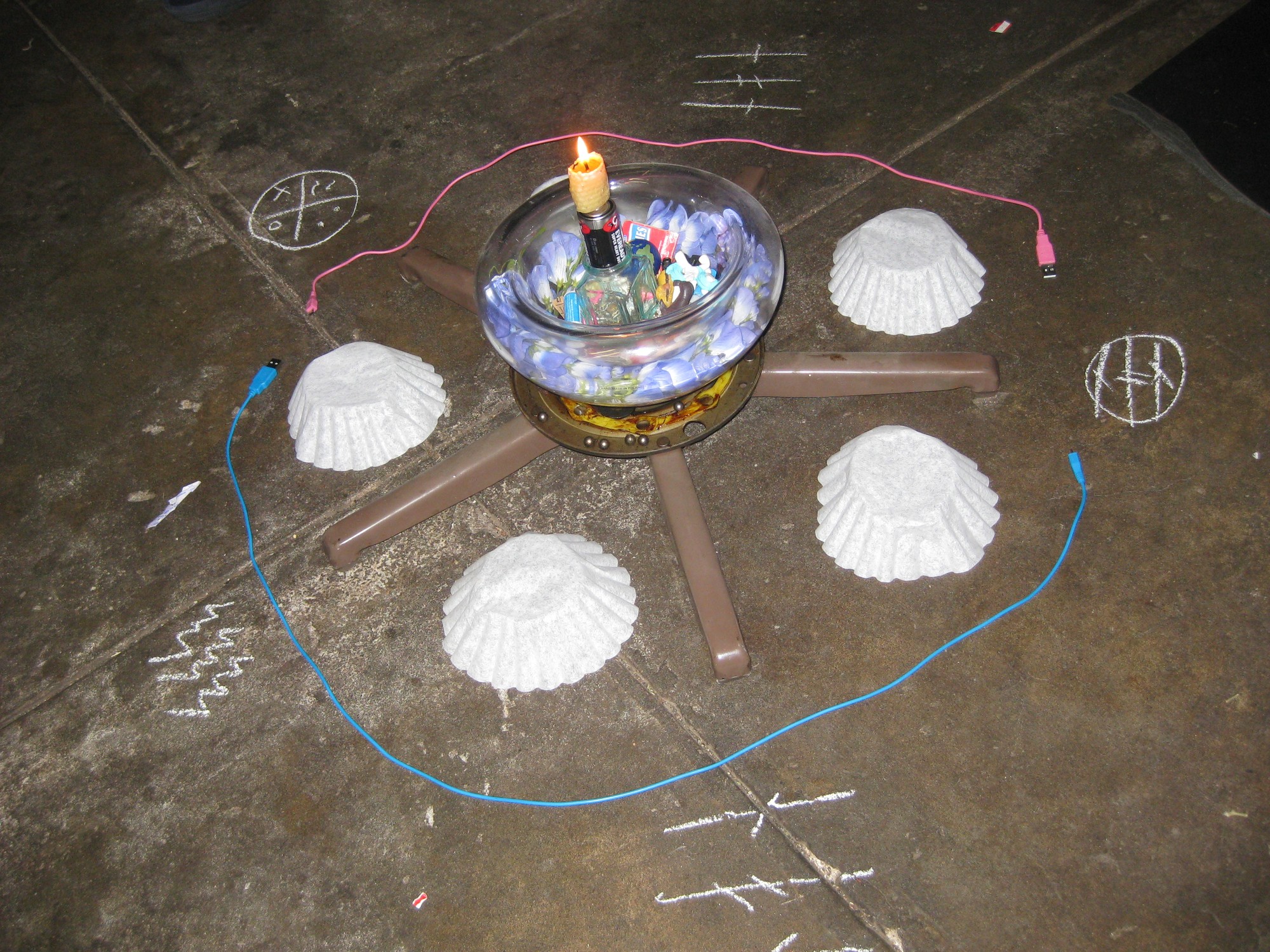 A metal base with five legs sits on a concrete floor circled by computer cables and symbols drawn with chalk. Between each leg is an upside-down coffee filter. A transparent bowl on top of the base has small objects inside, including blue flowers and a lit beeswax candle on top of a battery on top of a plastic ice cube.