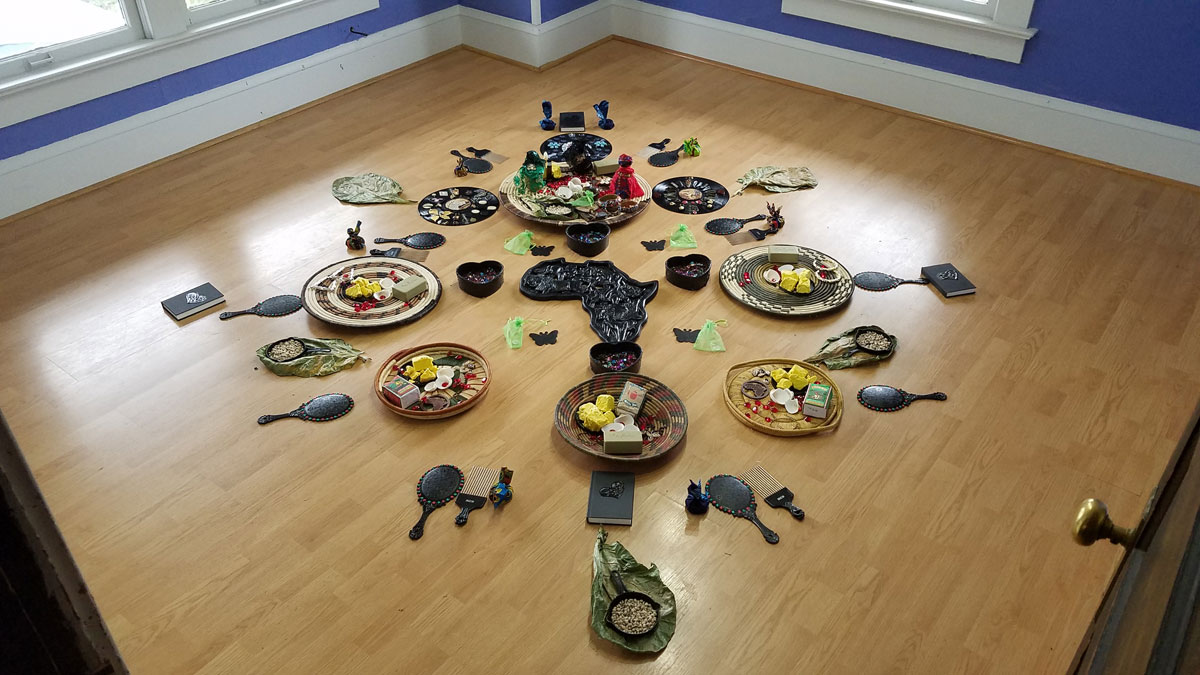 A variety of objects arranged in a symmetrical pattern on the floor around a wood carving in the shape of the African continent.