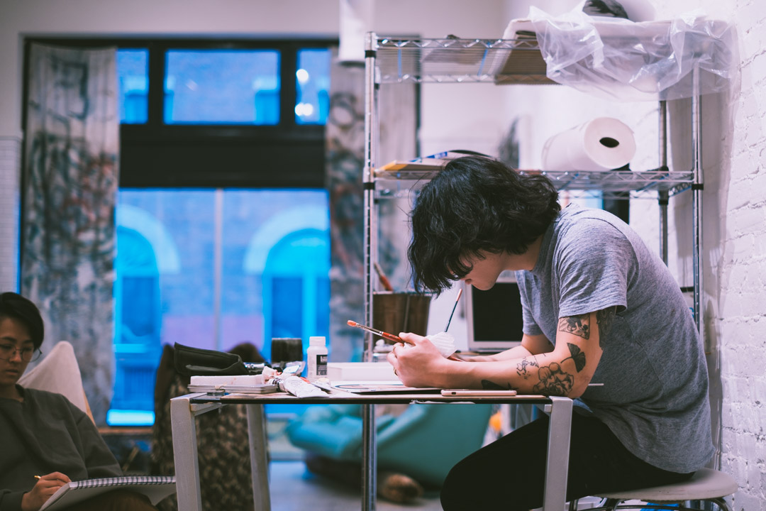 A person leans over a desk painting while another person sits drawing. They are in a room with a white-painted brick wall, art supplies on shelves and a large window.