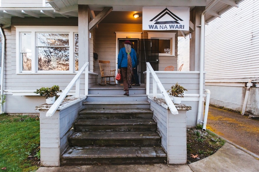 A person walks up the steps of an early twentieth-century home, a banner hung from the porch displays the Wa Na Wari logo.